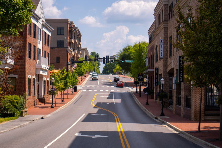 The view down a street in Downtown Blacksburg, Virginia, on a sunny summer day. The streets and brick sidewalks are neat and clean, the buildings are made of brick, and there are quaint street lamps along the road.