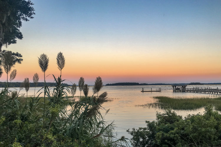 View from the river’s edge of a sunset over May River in Bluffton, South Carolina. Tall grasses slightly obscure the view, and a long dock reaches out into the water in the distance. 