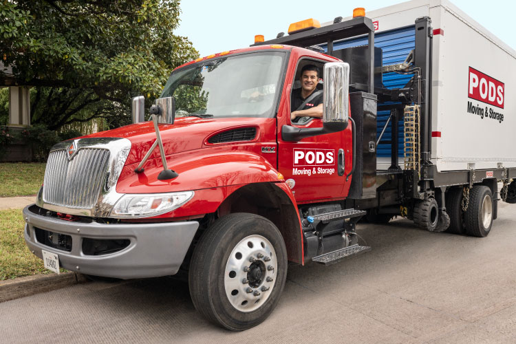 A PODS driver is smiling from the cab of his truck. He’s parked in front of a residential home, and there’s a PODS portable moving and storage container on the back of the truck. 
