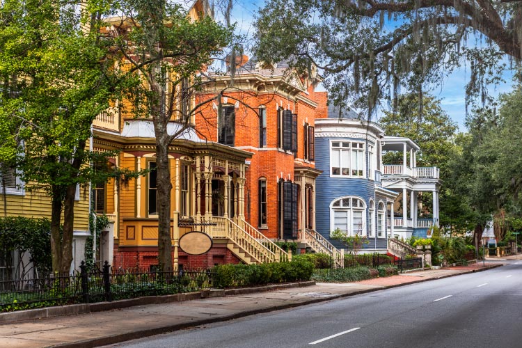 A row of colorful historic homes along Whitaker Street in Savannah, Georgia. The houses are each at least two stories tall and feature covered porches and ornate exterior design elements.