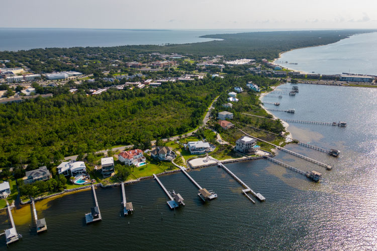 An aerial view of waterfront homes in Gulf Breeze, Florida. There’s a lush green wooded area behind the row of homes and, in front of nearly every house, there’s a long dock with a small boat house.