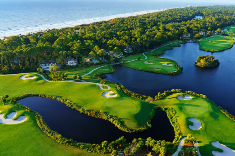Aerial view of a golf course in Hilton Head Island, South Carolina. There is a densely wooded area between the golf course and the beach. Luxury homes line the edges of the golf course.