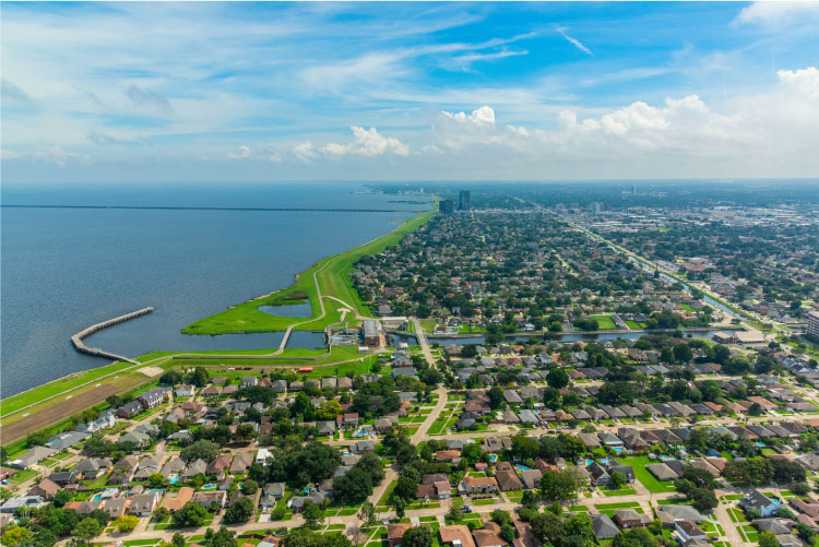 Aerial view of Metairie, Louisiana, on a summer day. The coastal city is sprawled out across flat terrain. A long causeway can be seen in the distance.