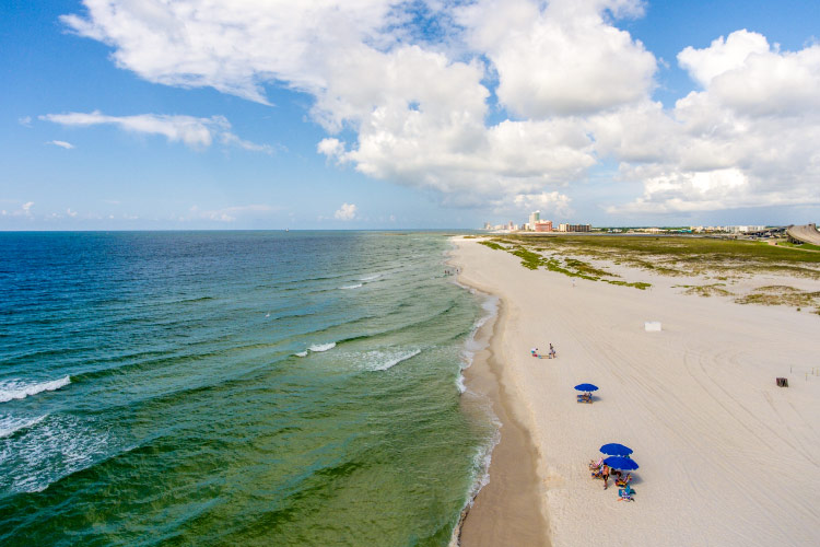 Aerial view of Orange Beach in Alabama. A few blue beach umbrellas are staked in the sand as locals enjoy a sunny day at the beach. 