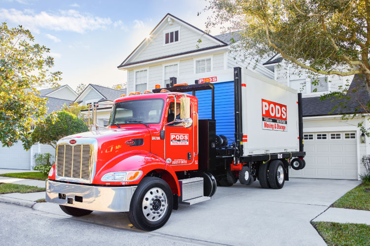 A PODS truck is backed into a residential driveway. There’s a PODS portable moving container on the bed of the truck, and the driver is preparing to use PODZILLA to position the container in the driveway so it can be loaded.