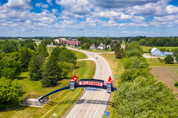 Ariel view of the welcome sign in Frankenmuth, Michigan, on a sunny day. The road is winding through a wooded area and in the distance, the town can be seen.