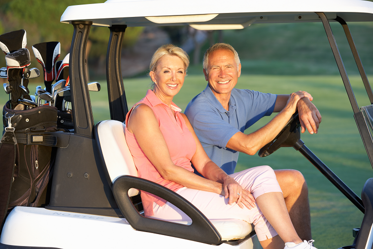 A mature couple smiles as they sit in their golf cart. Their caddies with clubs are in the back of the cart. The course can be seen in the background. 