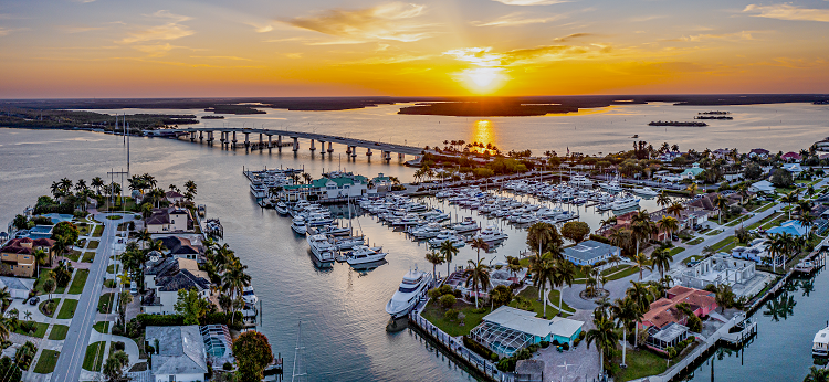 A panoramic of Marco Island, Florida. You can see a marina with various types of boats as well as luxury homes. There is a bridge, and the sun is setting. 