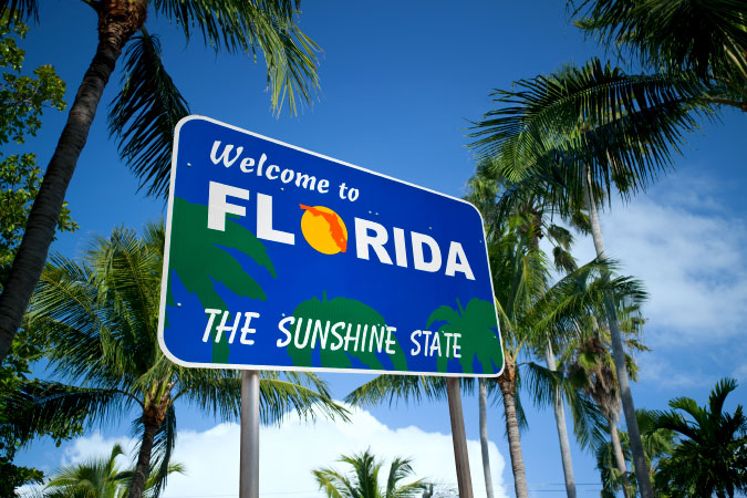 Street-level view of a “Welcome to Florida” interstate sign surrounded by palm trees on a sunny day.