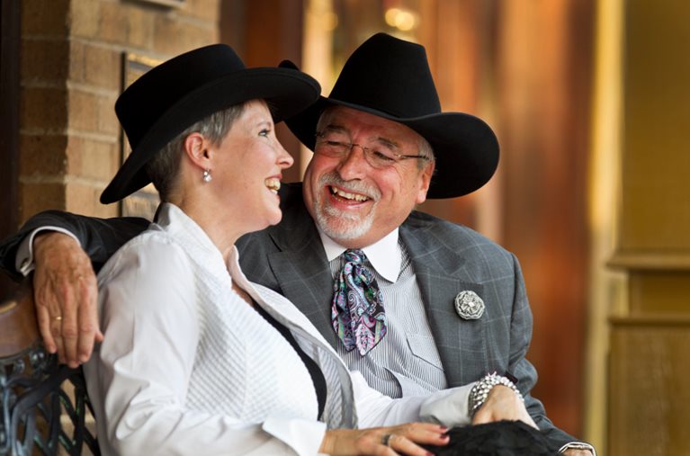 A happy retired couple is sitting together on a city bench in Texas. They’re dressed up in black western-style hats.
