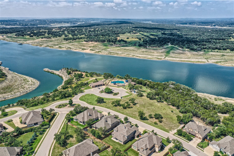 Aerial view of large residential homes in Lago Vista, Texas, beside Lake Travis. The houses appear similar, with gray shingle roofs and large floor plans. There’s a community pool along the bank of Lake Travis and a densely wooded area nearby.