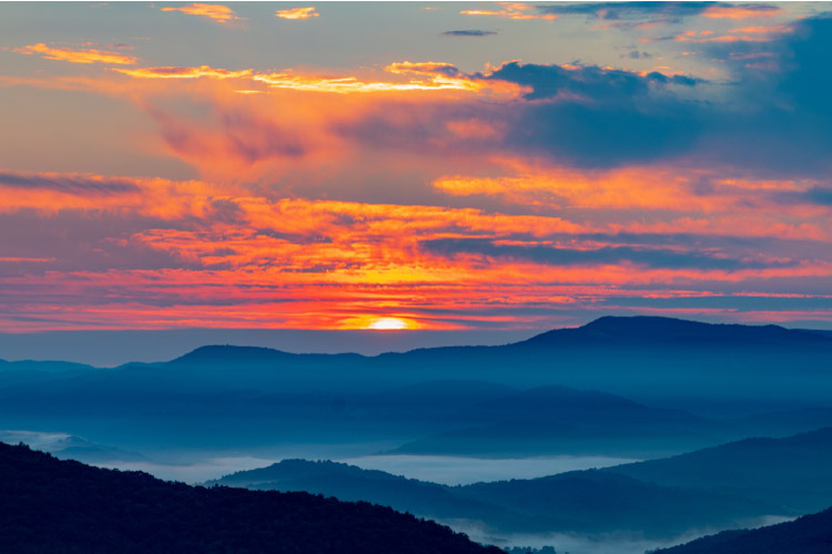 Gorgeous view of the Blueridge Mountains in Virginia during sunrise. A dense fog is settled between the rolling mountains, and the sky is a fiery pink and orange.