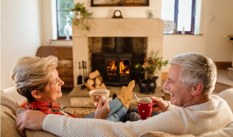 A mature couple is sitting on their couch in front of a roaring fire, drinking hot cocoa and laughing together. 