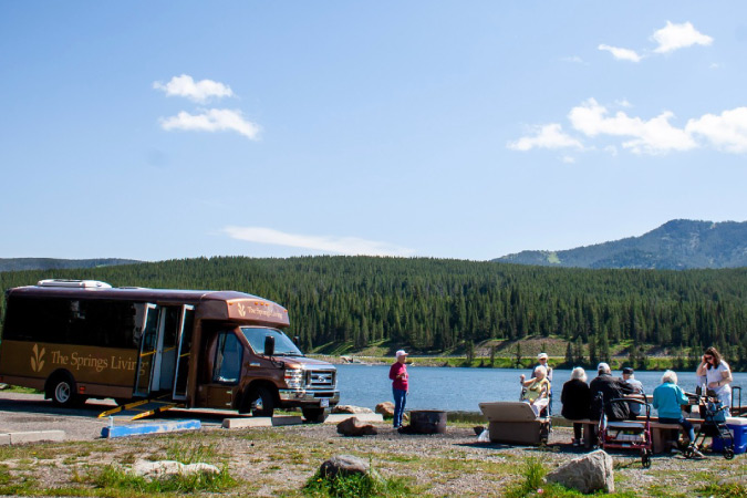 A large community van from The Springs at Bozeman retirement community is parked beside a river as  several residents enjoy the view from a rest area. 