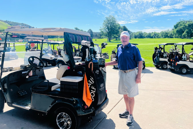 A resident of Mackenzie Place - Colorado Springs is posing for a photo by his golf cart at the Colorado Springs Country Club. 