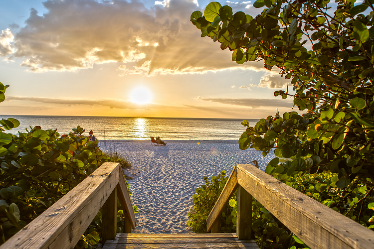 An image of the beach in Naples, Florida. The sand is white and there are two chairs with people sitting in them in the distance. The sun sets into the clouds on the image’s horizon. 