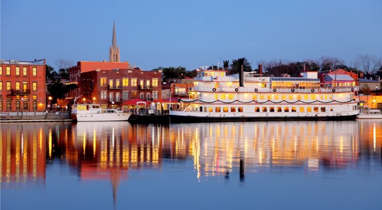 View of Wilmington, NC, after sunset from across the Cape Fear River. There’s a river boat anchored at the river's edge and a church steeple rising up in the distance.