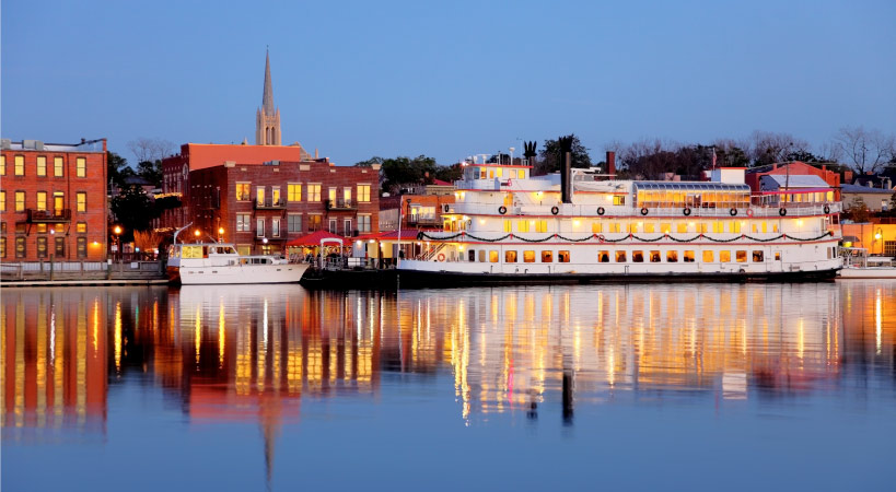 View of Wilmington, NC, after sunset from across the Cape Fear River. There’s a river boat anchored at the river's edge and a church steeple rising up in the distance.  