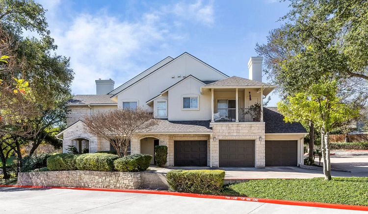 A white and tan multi-story home in the Allandale neighborhood of Austin, TX. It is surrounded by green trees and shrubs as well as a low brick wall. 