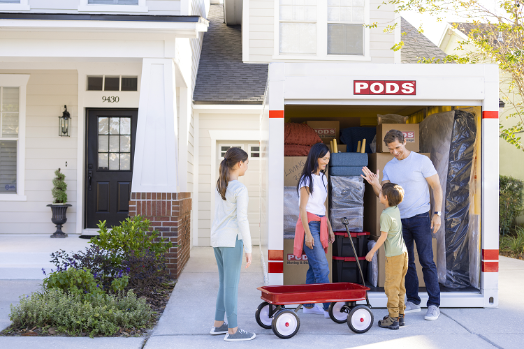 A family of four celebrates packing their fully loaded PODS container as they get ready to move to Austin. The container sits in their driveway and there is a red wagon in the frame. 