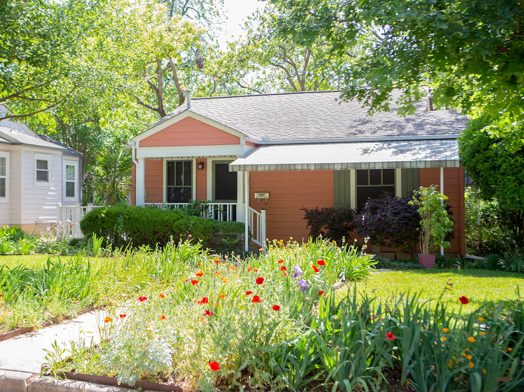 A red one-story home in the Rosedale neighborhood of Austin, Texas. It sits in the shade, covered by tall green trees. 