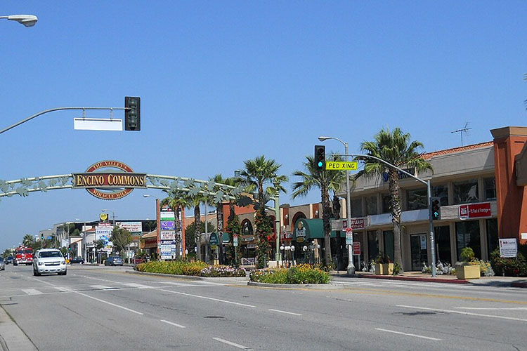 An image of downtown Encino in Los Angeles. There is a strip mall with stores and a sign that reads “Encino Commons.” There are also cars driving through. 