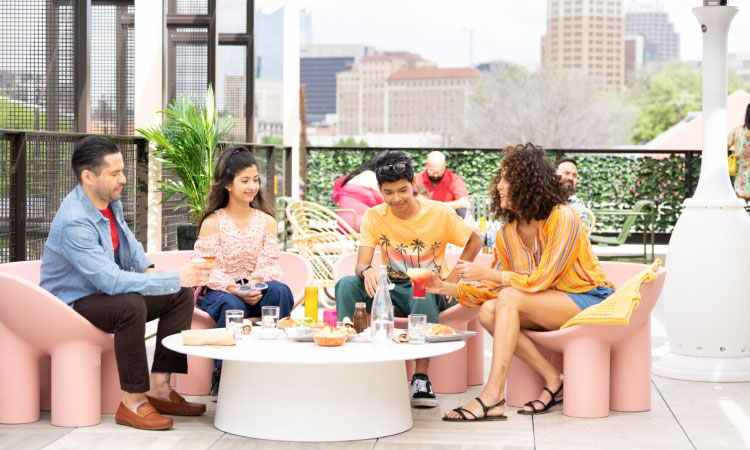 Friends are enjoying brunch at a rooftop restaurant in San Antonio, Texas, on a sunny autumn day. The furniture is eclectic, with exaggerated chair legs and low chair backs. You can see the city skyline in the distance.