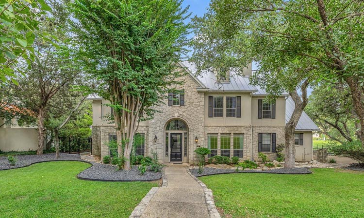 A luxury home in the North Central area of San Antonio. There is a wide walkway leading up to the front door through a nearly kempt lawn. The house is two stories with a stone facade front and a metal roof.