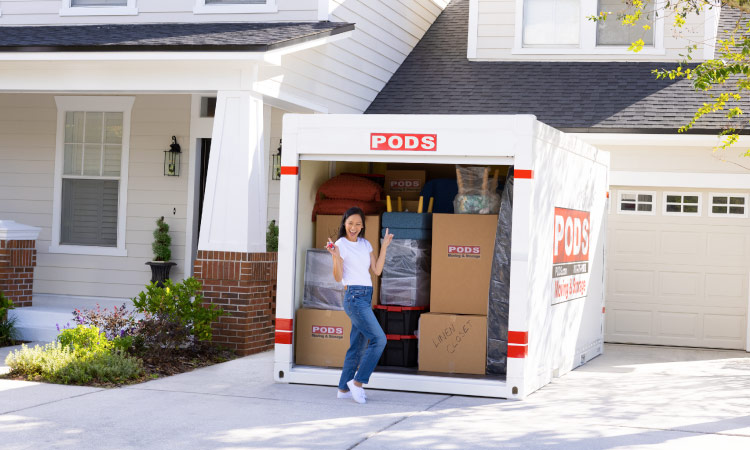 A woman is striking an excited pose in front of her loaded PODS container. The container is in her driveway and its door is open. The inside of the container is loaded with boxes and wrapped furniture, ready to be transported to her new home in San Antonio, Texas.