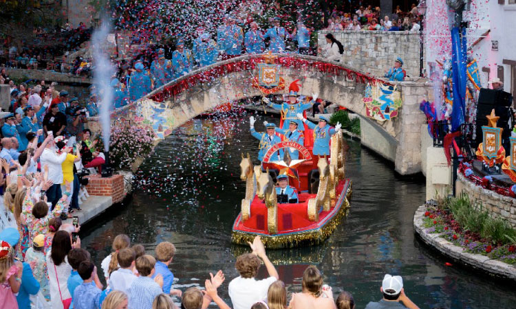 A red and gold boat filled with Texas Cavaliers cruises beside the San Antonio River Walk during the Texas Cavaliers River Parade, a part of the Fiesta San Antonio celebrations. Onlookers cheer and toss confetti into the air as the boat passes by.