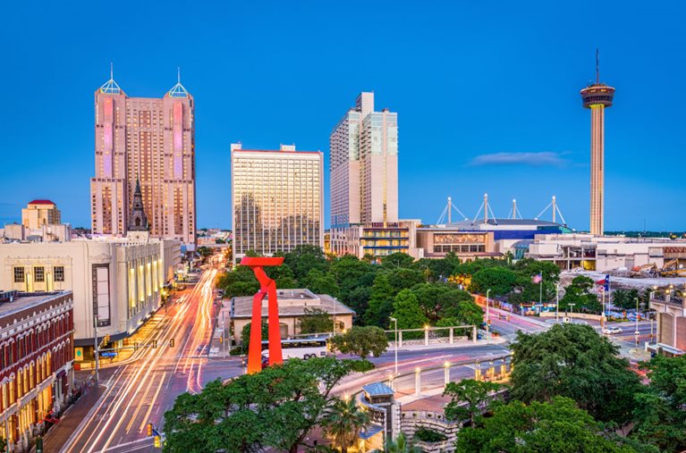 The San Antonio, Texas, skyline at dusk. Streams of light illuminate the streets of the city as cars travel through the avenues. A few tall buildings break up the clear blue of the sky and a large red sculpture decorates the center of the image.
