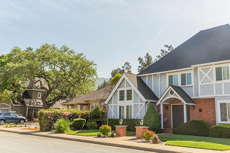 A two-story light blue Bavarian-style home in Almaden Valley in San Jose, California. There is brick around the door with tall hedges and bushes in a green yard. 