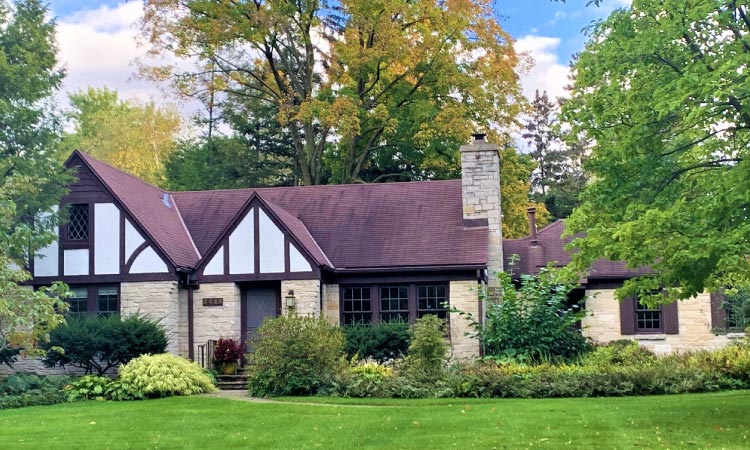 A large, Tudor-style home in the Madison, Wisconsin, suburb of Shorewood Hills. The home features classic Tudor detailing, a chimney, and a stone facade. The yard is kempt, and there are various trees and shrubs decorating the front of the home.