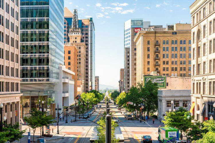 A view down a main thoroughfare in Downtown Salt Lake City. The two directions of traffic are separated by a lush, green-filled median and bordered by large city buildings.