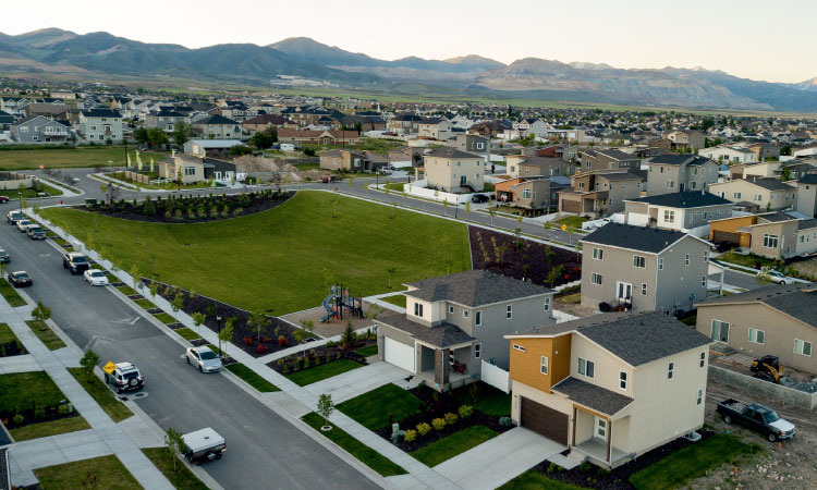 Aerial view of a contemporary residential area in Salt Lake City, featuring a mix of stylish single- and two-story homes. Majestic mountains form a scenic backdrop in the distance, while a serene green field stretches between some of the neatly laid out residential streets.