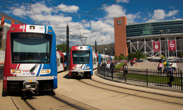 Two TRAX light rail trains are arriving at Rice-Eccles Stadium in Salt Lake City on a sunny day. Pedestrians are walking along a sidewalk beside an iron fence, with the stadium entrance visible in the background. It’s a vibrant urban scene capturing public transportation and the bustling atmosphere around the stadium.