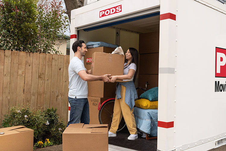 A young couple pack up their portable PODS moving and storage container as they move to Fort Worth. There are cardboard boxes everywhere, and the one behind the man reads “living room.” 