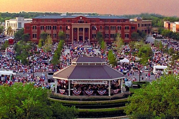 A panoramic aerial view of downtown Southlake, Texas. There is a crowd of people gathered in a park. There is a gazebo in the middle of the image. 