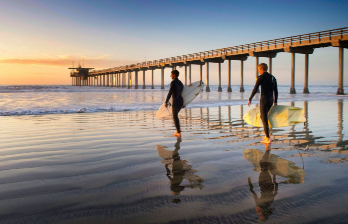 Two surfers walk across the glassy wet sand toward the water on a San Diego beach.