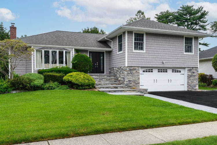 A split-level house in Syosset, New York, on Long Island. The house features a stone and shingle exterior, a two-car garage, and stone steps leading to the entrance. The front yard is landscaped with manicured shrubs and a lush green lawn.