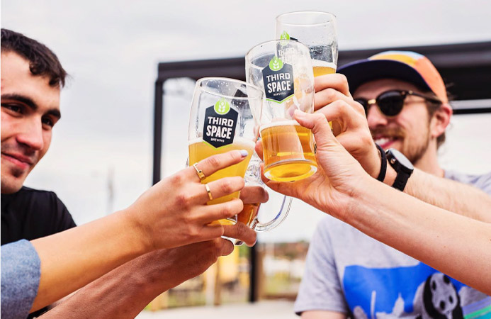 Four friends are clinking their beer glasses together at Third Space Brewing in Milwaukee, Wisconsin. Two of the friends’ faces are in frame and they’re smiling.