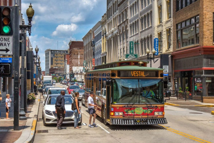 Three locals are lined up to board one of the free trolleys that operate in Knoxville, Tennessee. It’s a sunny day in the city, and the trolley is picking up passengers on a busy street.