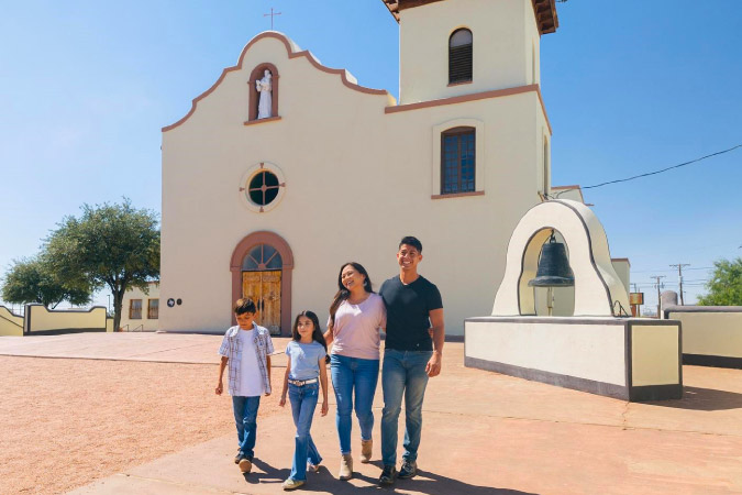 A family of four is walking near an adobe church in El Paso, Texas, on a sunny day.