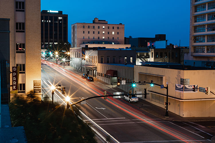 A street in downtown Tyler at dusk. Cars whizz by as lights shine through office windows from late-night workers.