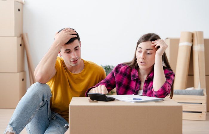 A young couple sits together beside a moving box, using a calculator, pen, and paper to calculate their interstate moving costs.