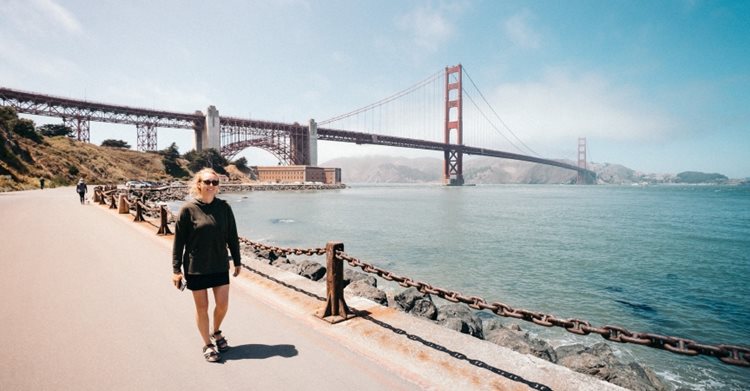 A woman is walking along the San Francisco coast on a sunny day with the Golden Gate Bridge in view behind her. 