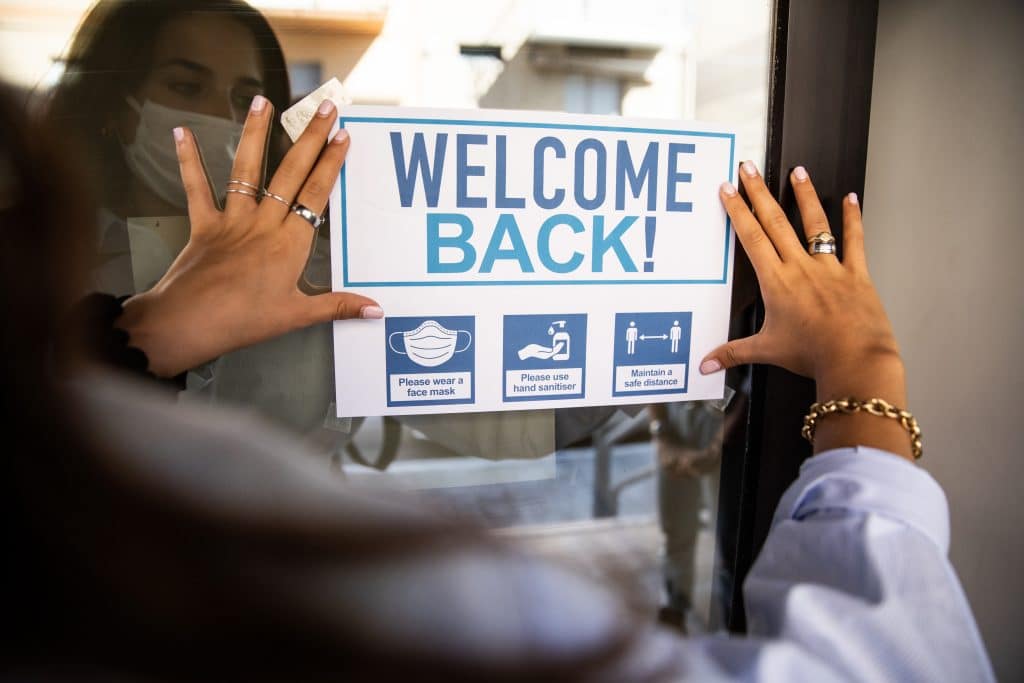 Female teacher wearing mask placing a "Welcome Back! sign on a campus door
